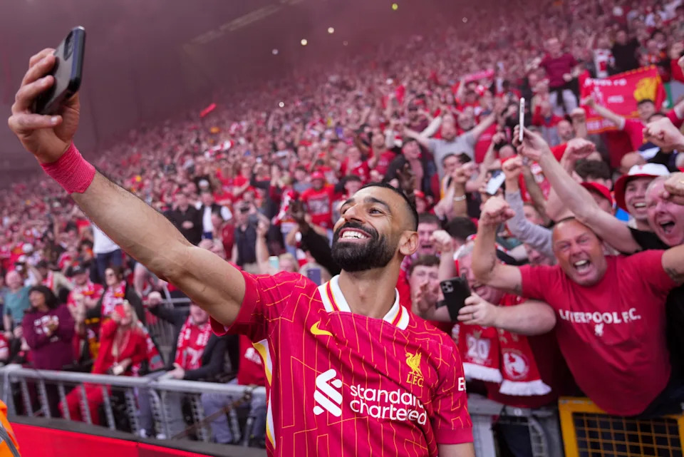 Mo Salah celebrating with Liverpool fans after scoring a crucial goal, holding a phone for a selfie.